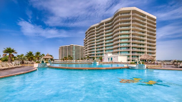 A hotel with a large swimming pool, palm trees, and a thatched-roof structure. - Beachview condo with lazy river, marina, multiple pools and internet (Orange Beach)