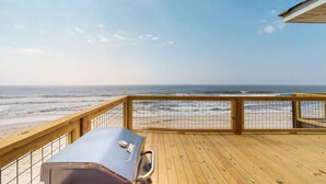Une terrasse en bois avec un barbecue, offrant une vue sur une plage et l’océan.