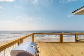 Une terrasse en bois avec un barbecue, offrant une vue sur une plage et l’océan.