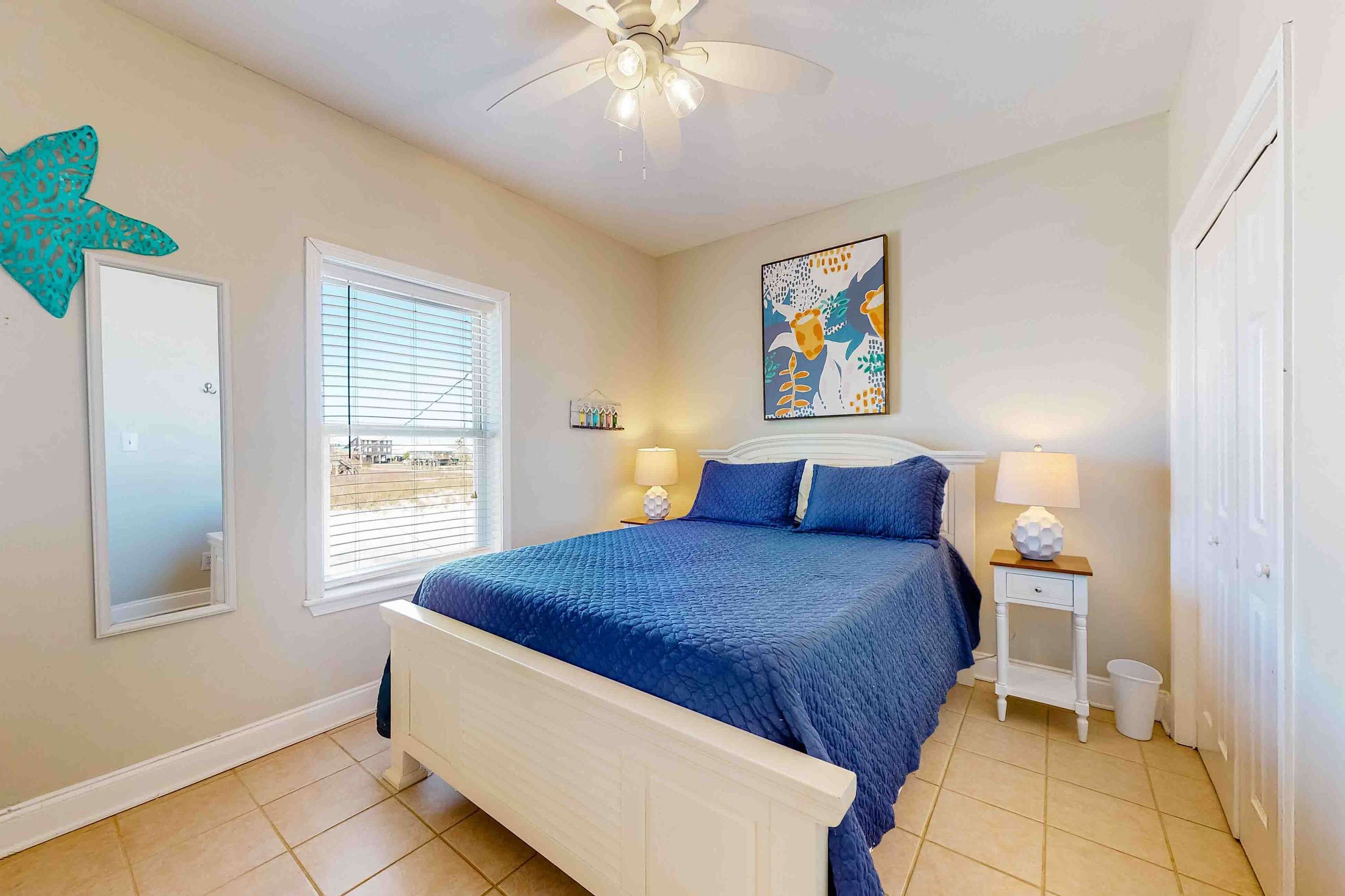 A bedroom with a bed, a ceiling fan, a window with blinds, a wall-mounted artwork, and a nightstand with a lamp.