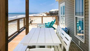 Una terraza de madera con una mesa y sillas blancas, con vista a una playa y una valla de madera.