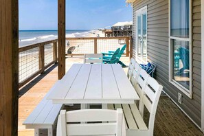 Une terrasse en bois avec une table et des chaises blanches, donnant sur une plage bordée d’une clôture en bois.