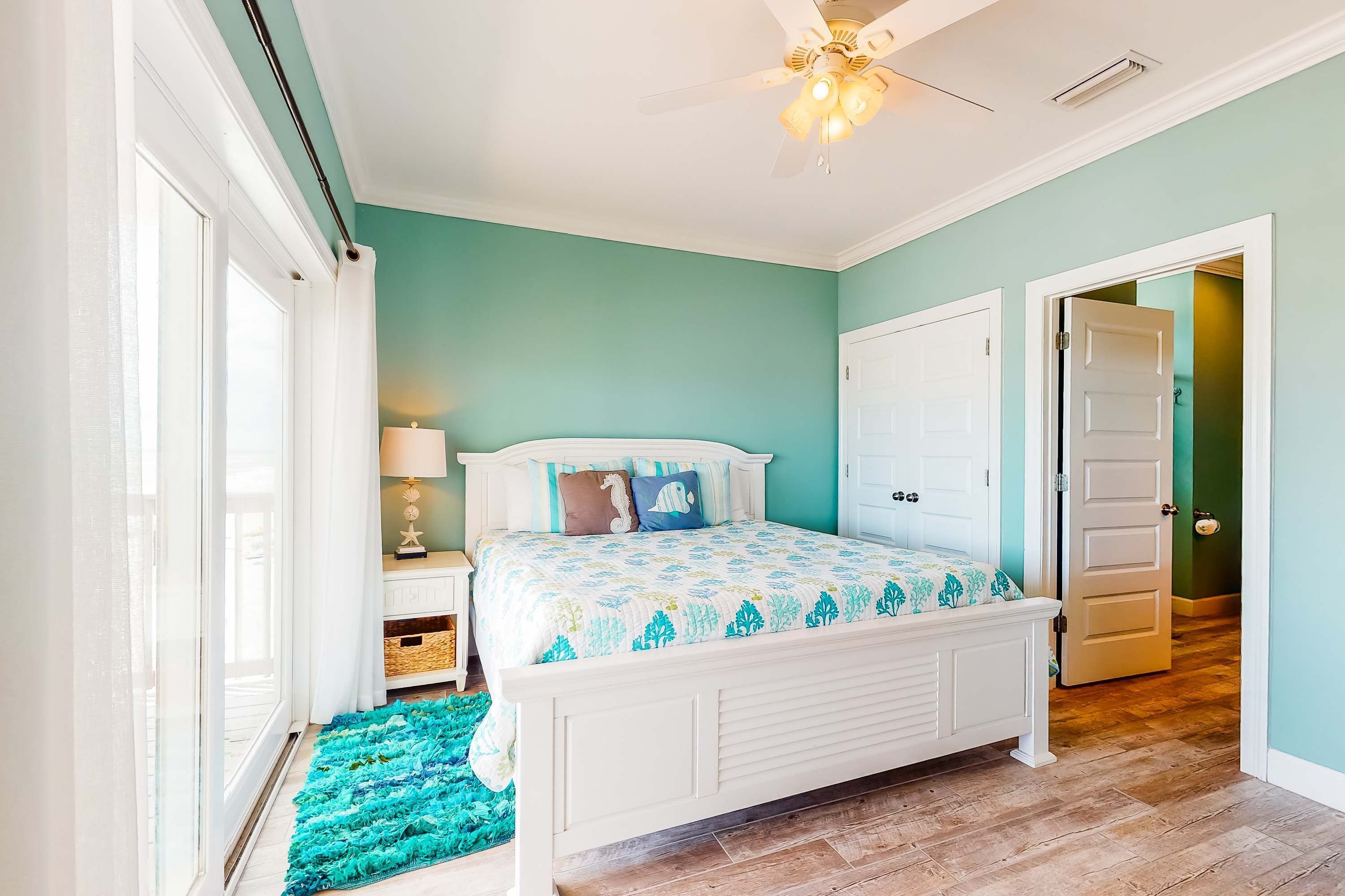 A bedroom with a bed, a nightstand, a lamp, a ceiling fan, and a door leading to another room.