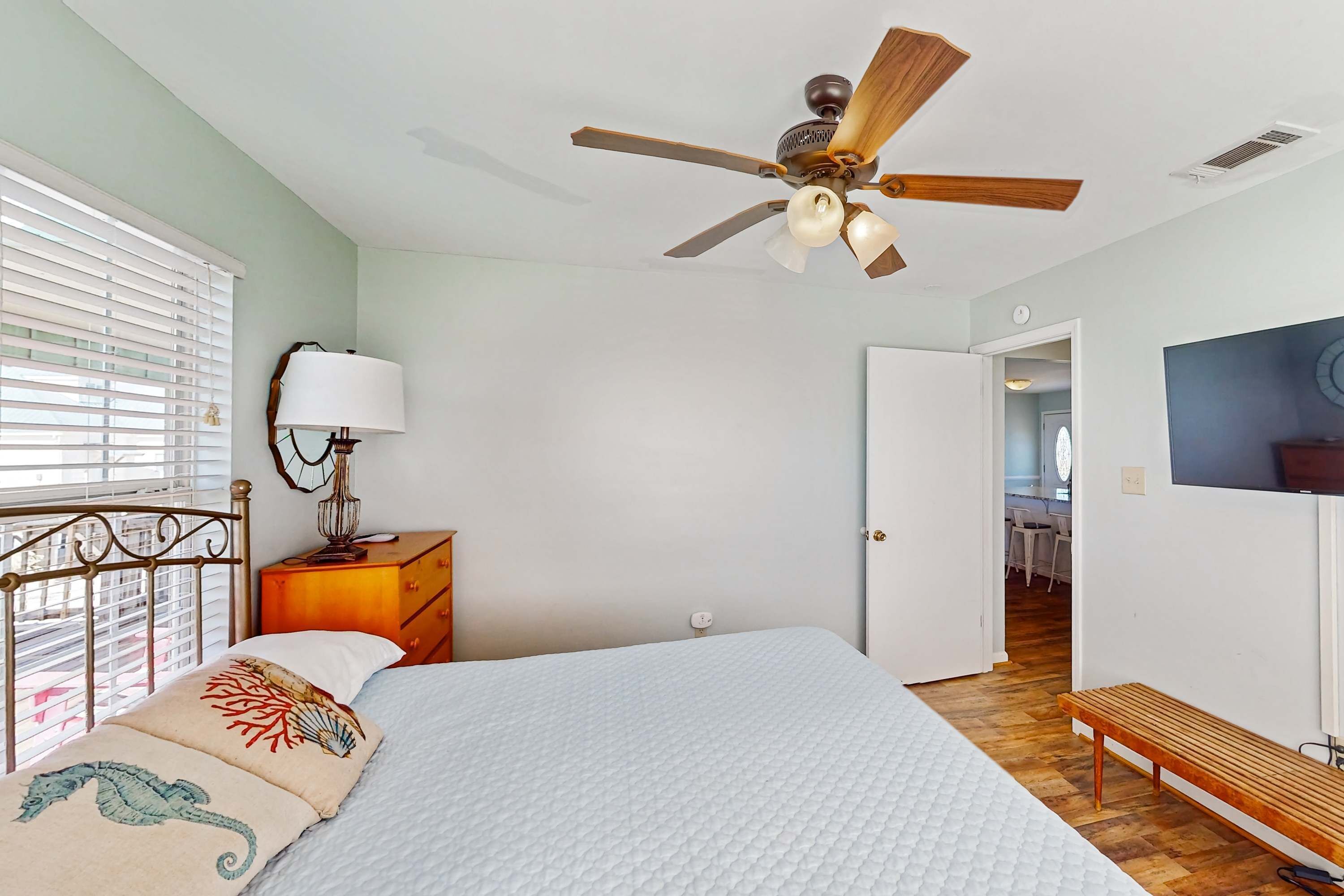 Une chambre à coucher comprenant un lit, un ventilateur de plafond, une table de chevet avec une lampe, un banc en bois et une porte donnant sur une autre pièce.