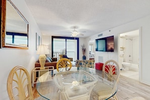 A dining area with a glass table and wicker chairs, a ceiling fan, and a view of the ocean through large windows. - Beachfront condo with private balcony, shared pool, and hot tub (Orange Beach)