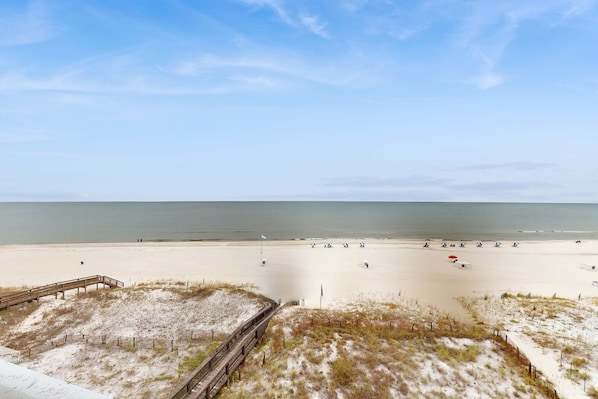 A beach with a wooden boardwalk leading to the shore, a flag, and a sandy area with umbrellas. - Beachfront condo with private balcony, shared pool, and hot tub (Orange Beach)