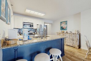 A kitchen with white cabinets, a blue island with stools, stainless steel appliances, and a wooden floor. - Beachfront condo with private balcony, shared pool, and hot tub (Orange Beach)