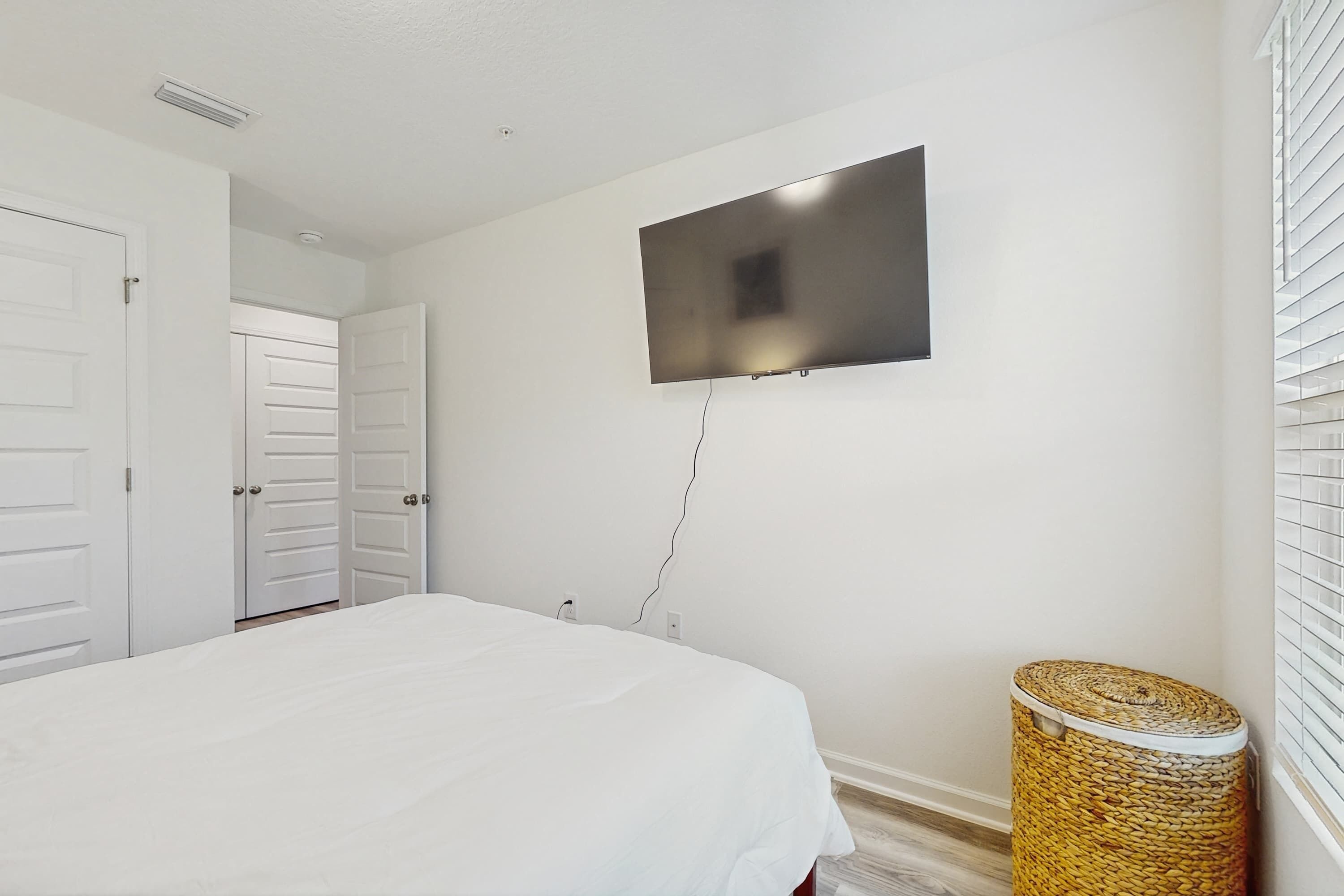 Een slaapkamer met een bed, een aan de muur bevestigde televisie, een rieten mand en een deur naar een andere kamer.