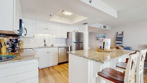 A modern kitchen with white cabinetry, a stainless steel refrigerator, and a central island with bar stools. - Beautiful beachfront condo with hot tub, pool, sauna, tennis, gym, & balcony (Orange Beach)