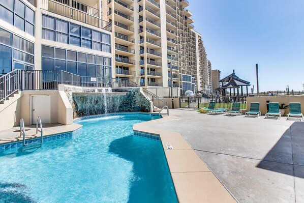 A swimming pool with steps leading into it, a gazebo, and lounge chairs on a sunny day. - Beautiful beachfront condo with hot tub, pool, sauna, tennis, gym, & balcony (Orange Beach)