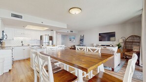 A modern kitchen with white cabinetry, a wooden dining table, and white chairs. There is a large window with a view of the outdoors. - Beautiful beachfront condo with hot tub, pool, sauna, tennis, gym, & balcony (Orange Beach)