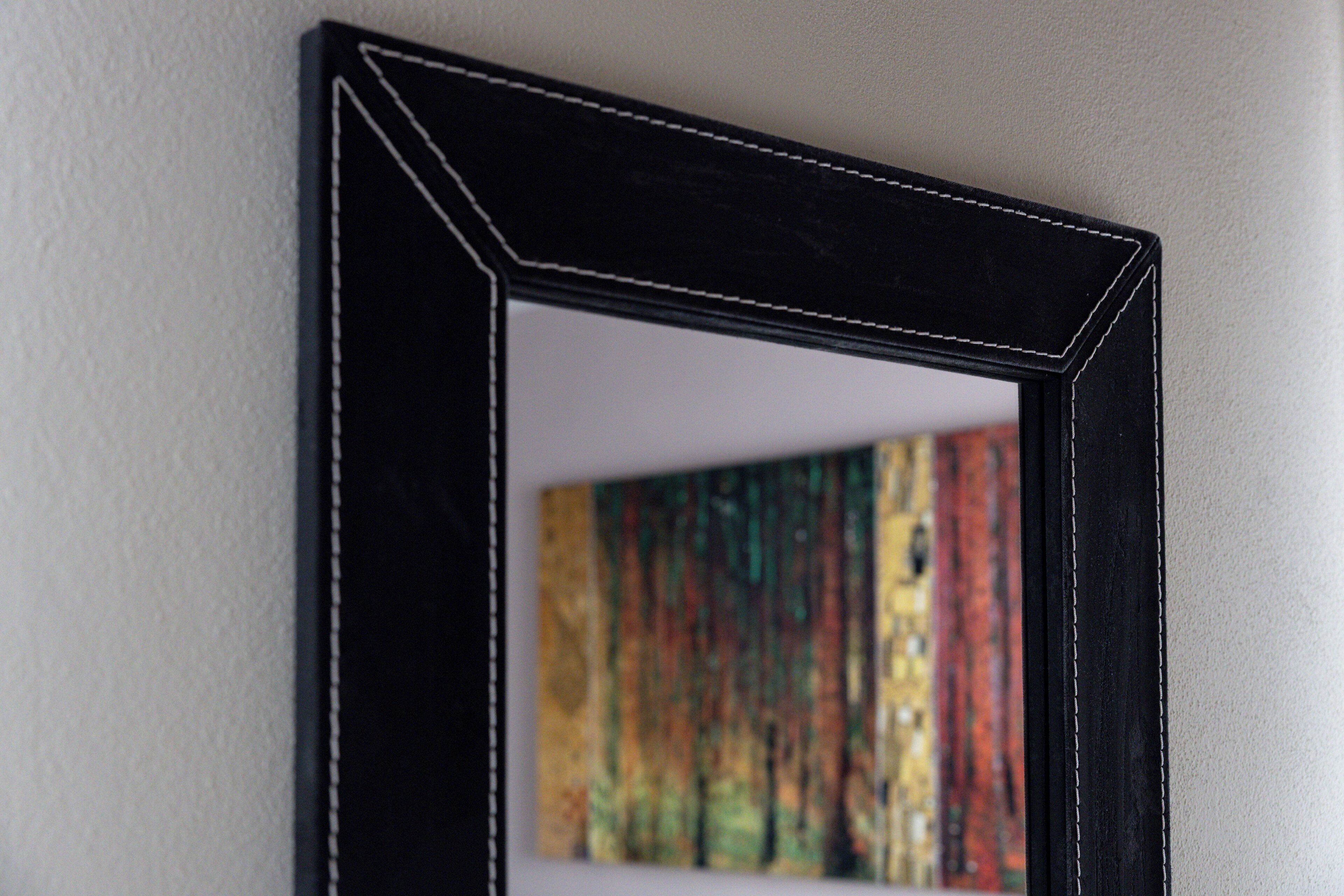 Ein Spiegel mit schwarzem Rahmen, der einen farbenfrohen Gemälde an der Wand reflektiert.