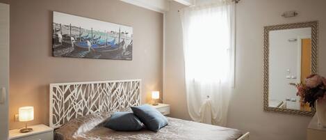 Un dormitorio con cama, mesitas de noche, un cuadro en la pared, un espejo y una ventana con cortinas.