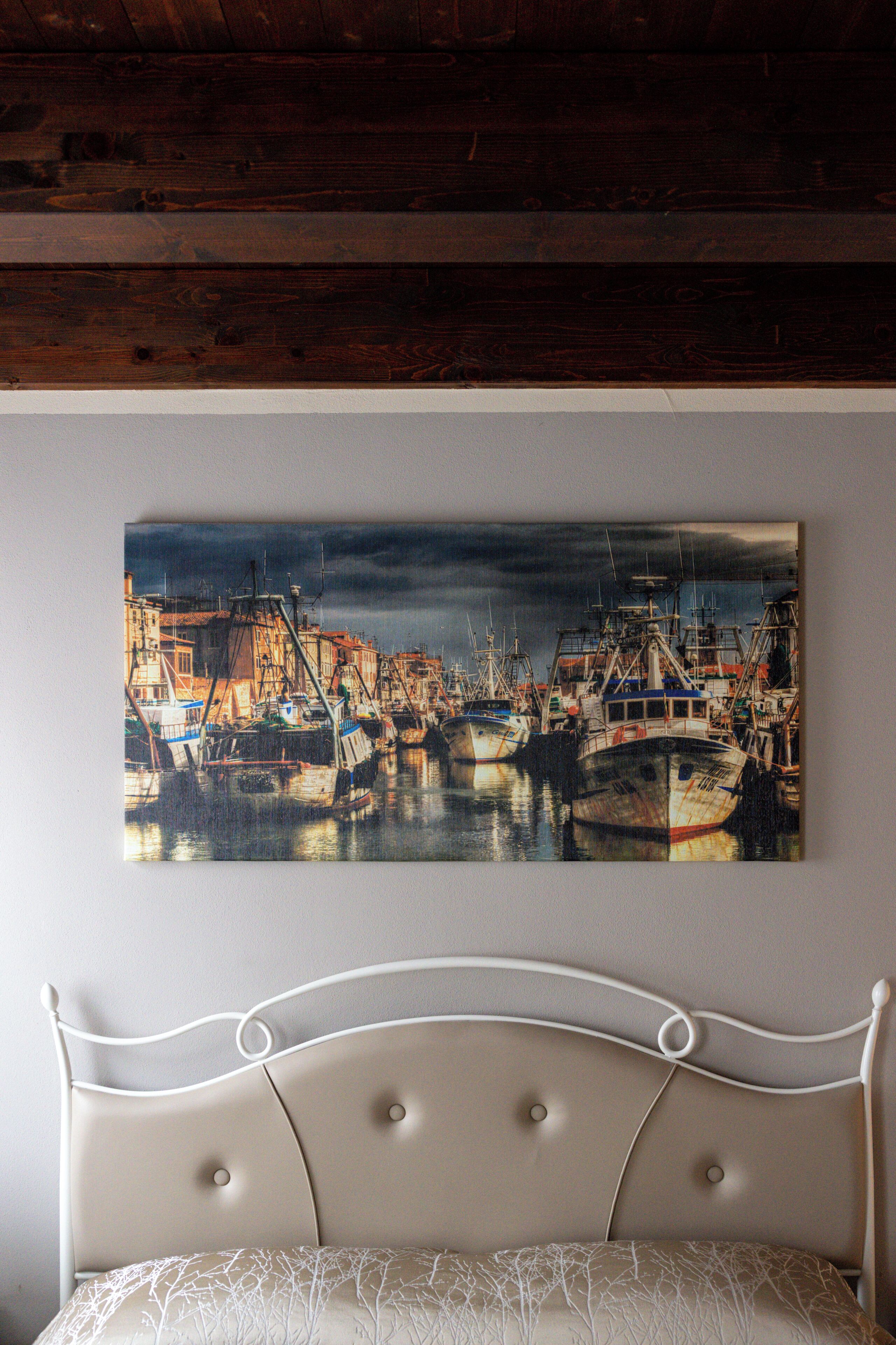 A bedroom with a bed, a wall-mounted painting of boats, and a wooden ceiling.