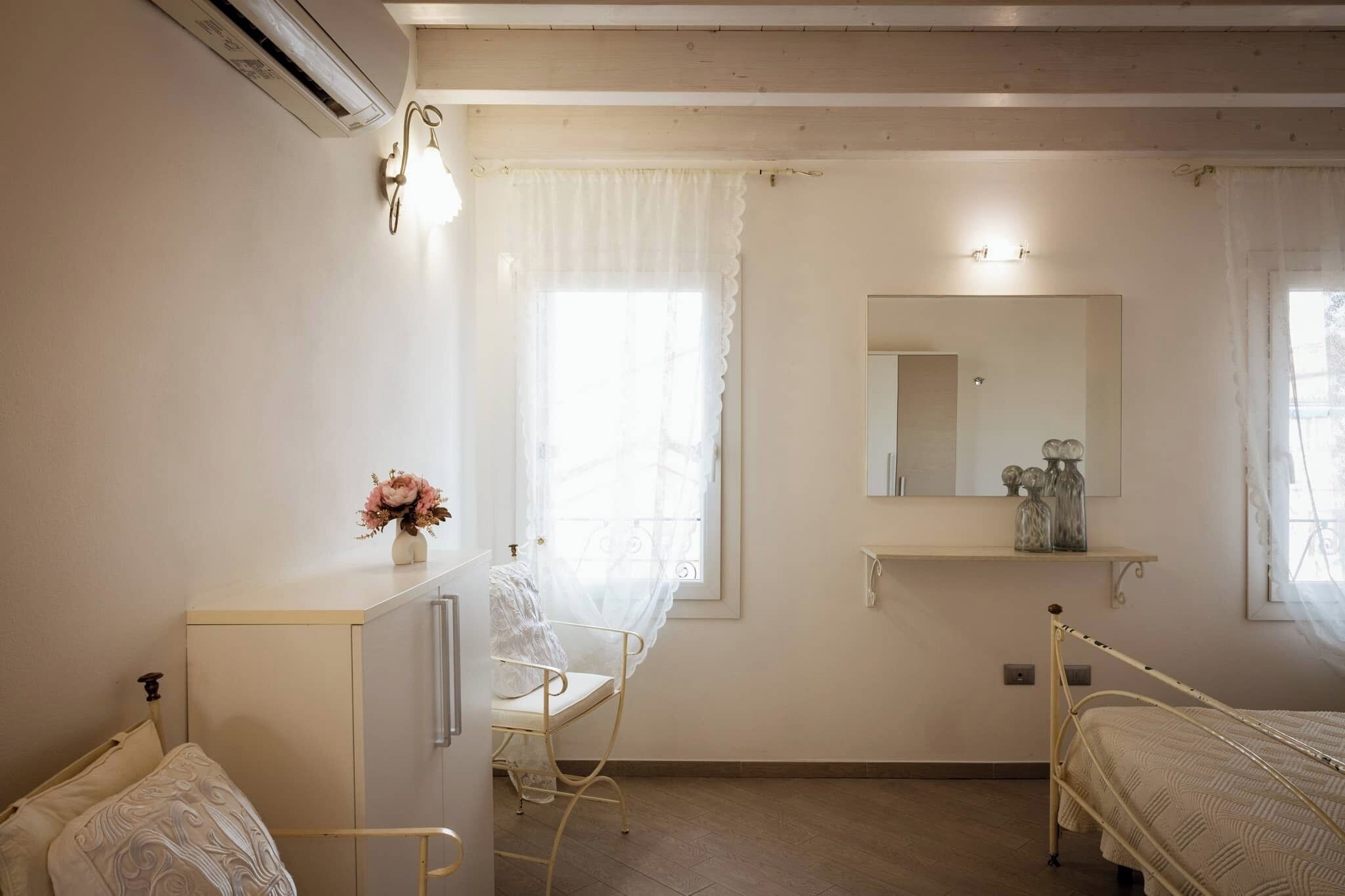 Um quarto com cama, mesa de cabeceira, cadeira, espelho e janela com cortinas.