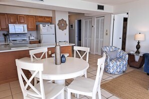Cocina compacta con armarios de madera, microondas y refrigerador. Hay una zona de comedor con una mesa redonda y sillas blancas. También se ve un sillón estampado y una mesita con una lámpara.