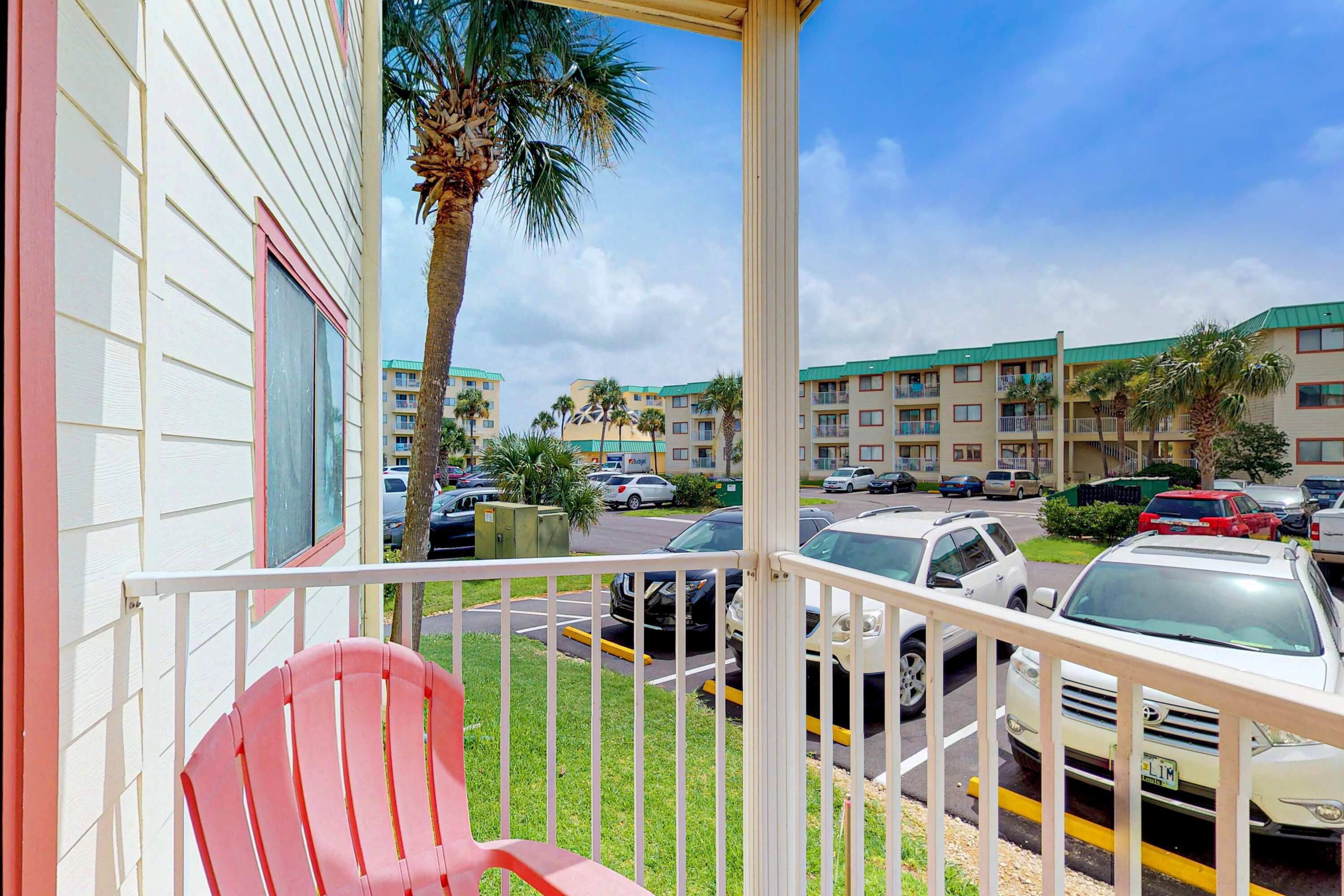A balcony with a red chair, a parking lot with several cars, and a row of multi-story buildings with green roofs in the background.