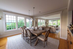 A spacious dining area with a wooden table and chairs, large windows offering a view of greenery, and a kitchen in the background.
