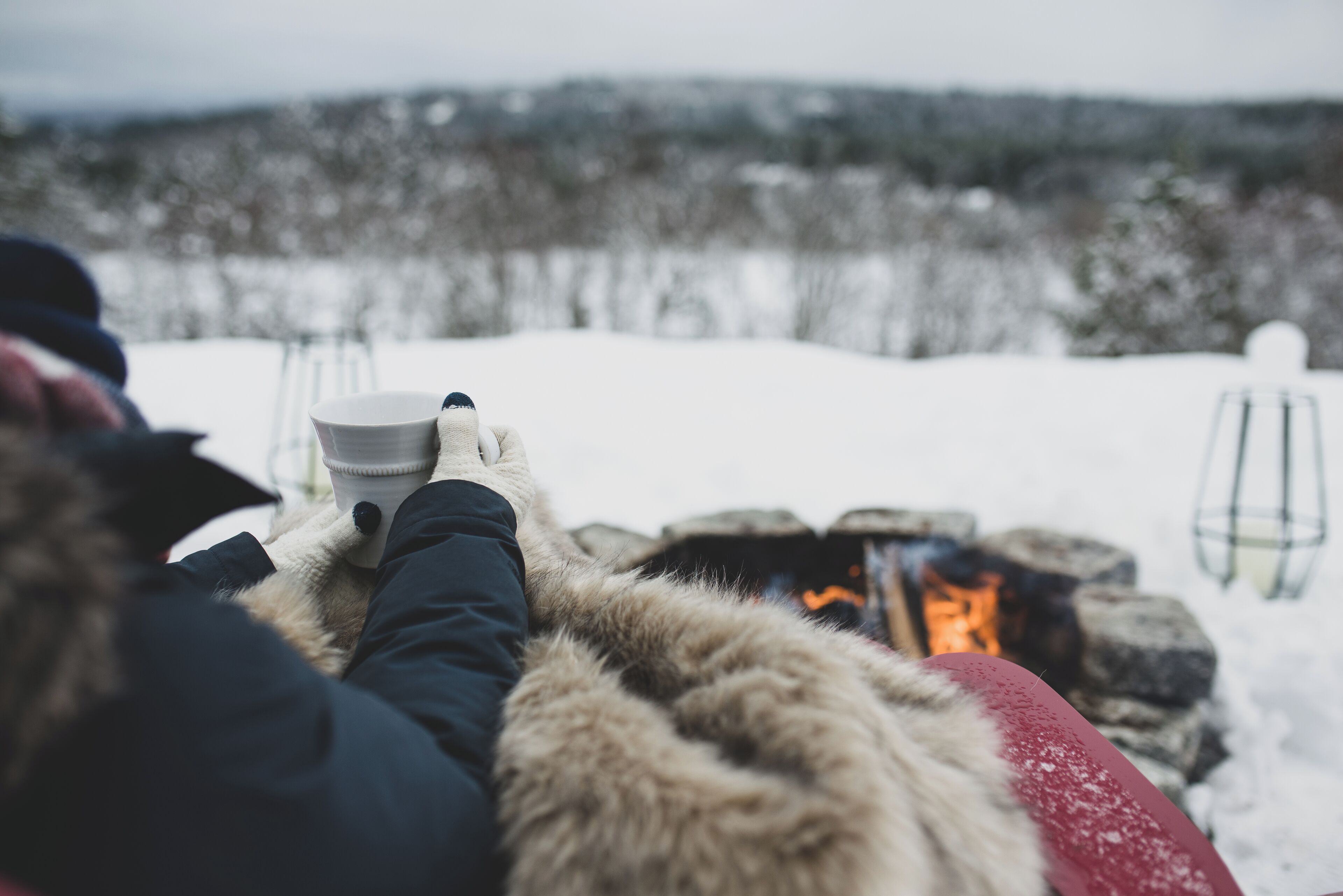 A person sitting by a fire, holding a cup, with a snowy landscape in the background.