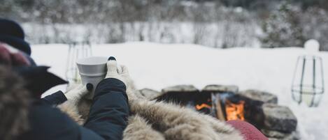 A person sitting by a fire, holding a cup, with a snowy landscape in the background.