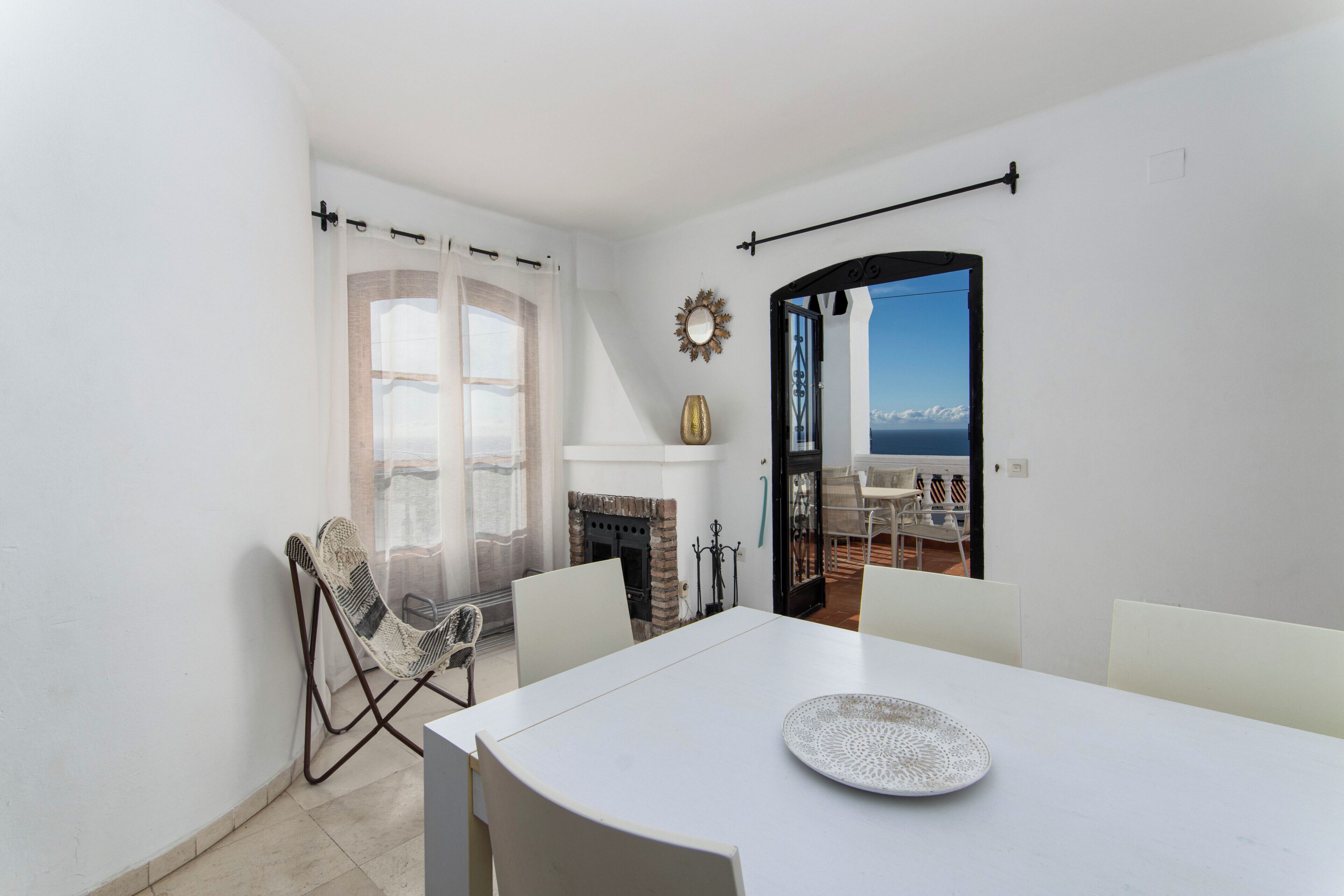 Een kamer met een witte tafel, een rieten stoel, een open haard en uitzicht op zee door een geopende deur.
