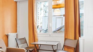A small room with a table and two chairs, a window with curtains, and a radiator. - Studio Raffine Quartier La Muette (Paris)