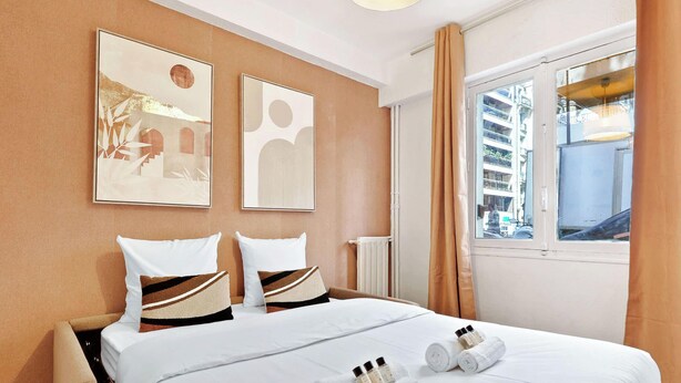 A hotel room with a bed, two pillows, a window with curtains, and two wall art pieces. - Studio Raffine Quartier La Muette (Paris)