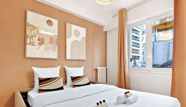 Ein Hotelzimmer mit einem Bett, zwei Kissen, einem Fenster mit Vorhängen und zwei Wandbildern.