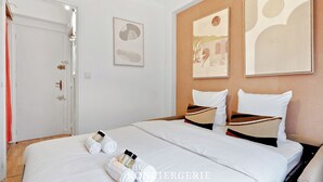 A hotel room with a neatly made bed, a door with a towel hanging on it, and two framed artworks on the wall. - Studio Raffine Quartier La Muette (Paris)
