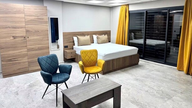 Moderni hotellihuone, jossa on sänky, kaksi tuolia, pieni pöytä ja suuri ikkuna, jossa on verhot.