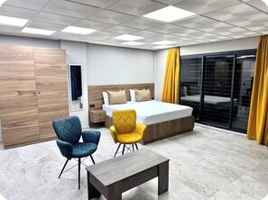 Une chambre d’hôtel moderne, équipée d’un lit, de deux fauteuils, d’une petite table et d’une grande fenêtre avec des rideaux.