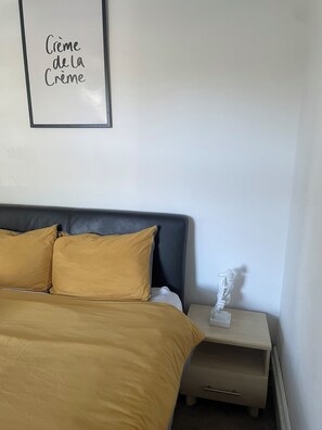 Ein ordentlich bezogenes Bett mit dunklem Kopfteil und zwei Kissen, ein Nachttisch mit einer Skulptur und ein gerahmtes Schild an der Wand.