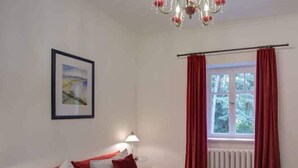 A bedroom with a bed, a nightstand, a lamp, a window with red curtains, and a chandelier. - Strandvilla Hubertus - H2 - Frida Kahlo (Ostseebad Boltenhagen)