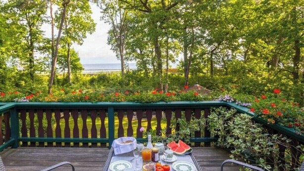 An outdoor dining area with a table set for breakfast, surrounded by lush greenery and a view of the sea. - Strandvilla Hubertus - H2 - Frida Kahlo (Ostseebad Boltenhagen)
