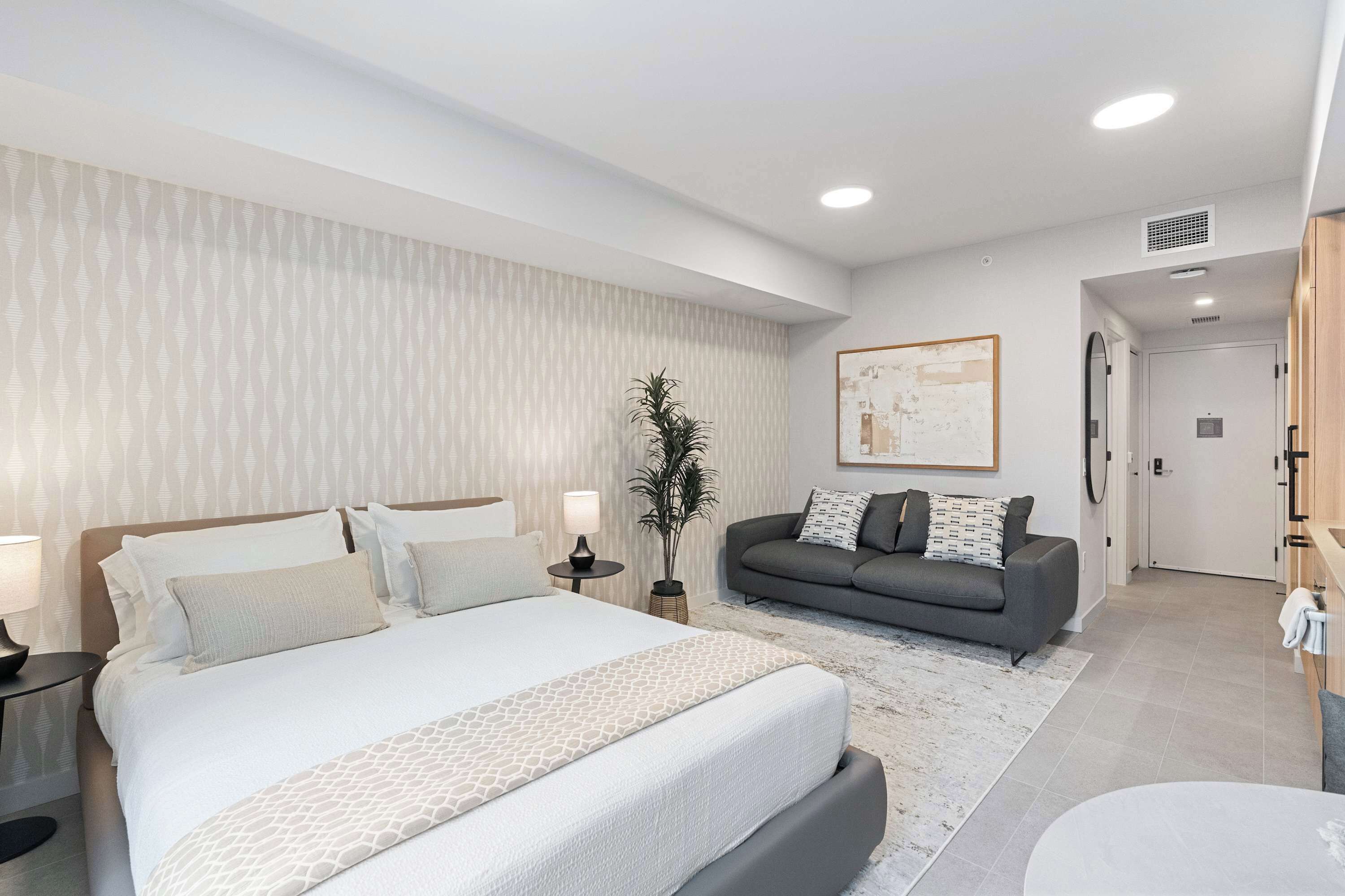 A modern hotel room with a large bed, a grey sofa, a small table with a lamp, and a plant.