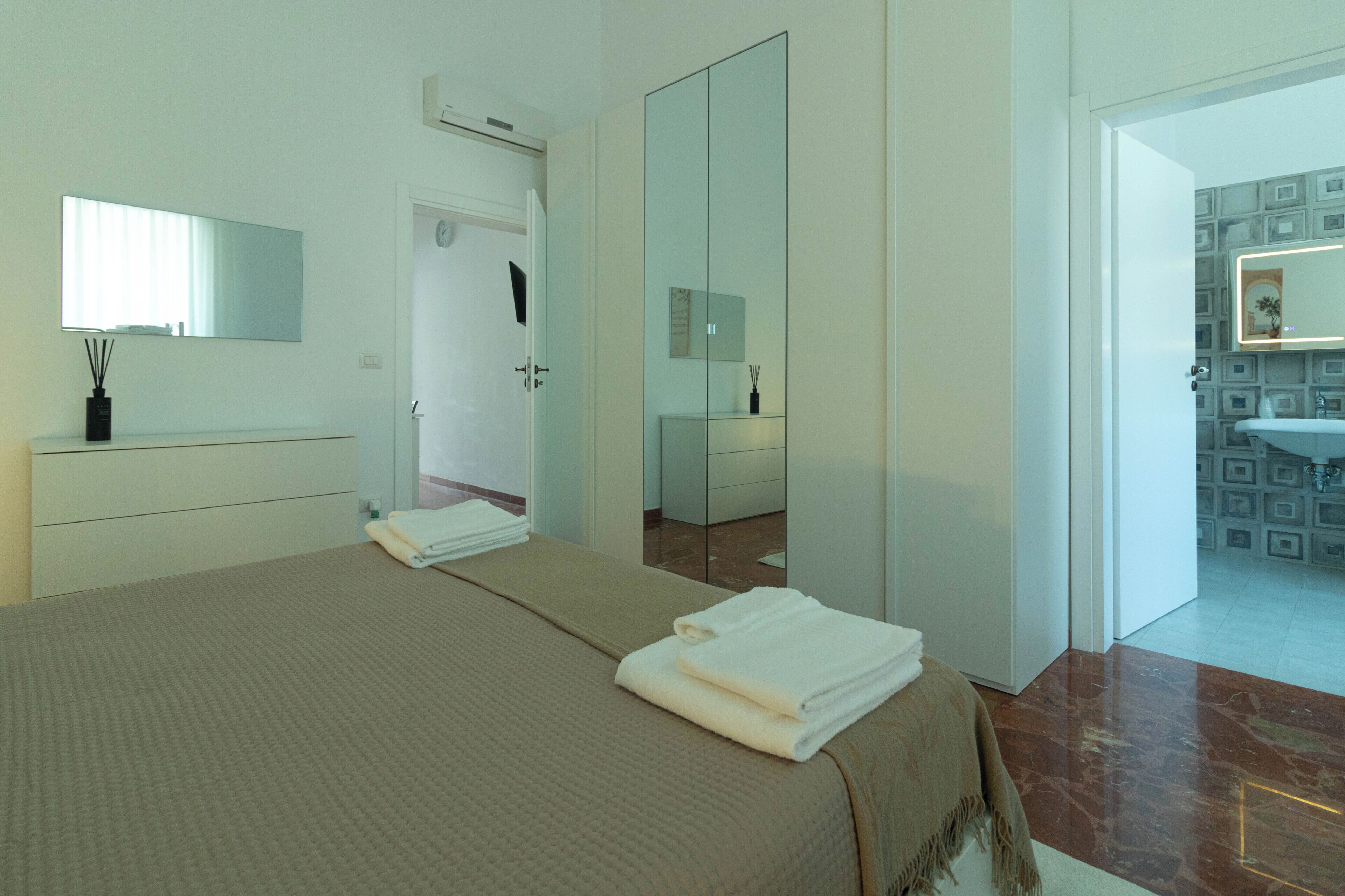 Une chambre d’hôtel avec un lit, une table de chevet, un miroir et une salle de bain visible à travers une porte ouverte.