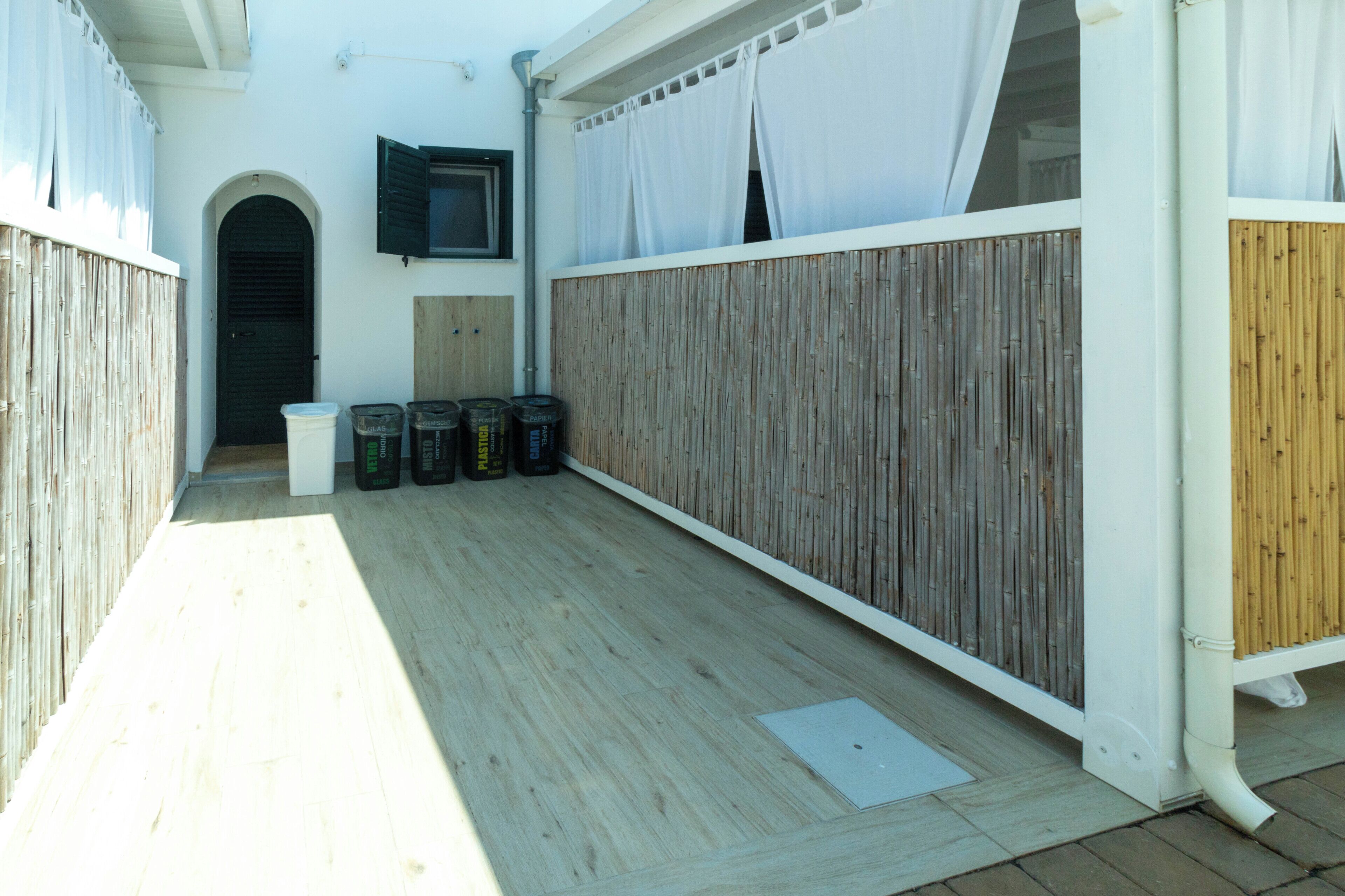 Ein überdachter Patio-Bereich mit Holzboden, einer weißen Wand, einer schwarzen Tür und einem Fenster mit Vorhang.