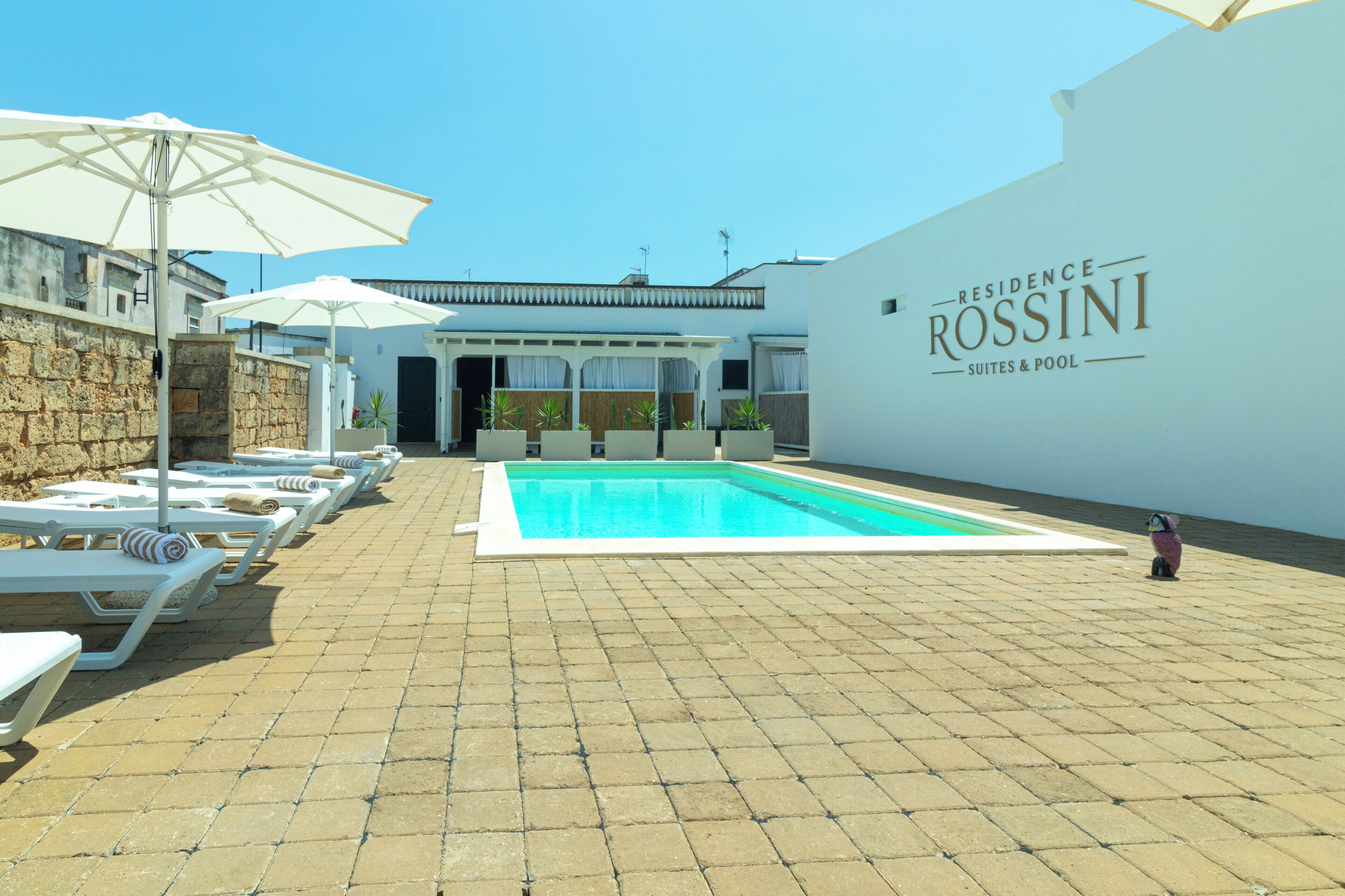 A pool area with lounge chairs and umbrellas, a clear sky, and a building with 'Residence Rossini Suites & Pool' signage.