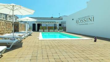 A pool area with lounge chairs and umbrellas, a clear sky, and a building with 'Residence Rossini Suites & Pool' signage.