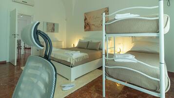 A bunk bed with white bedding, a white chair, a bed with a beige blanket, a small table with a lamp, and a door leading to another room.