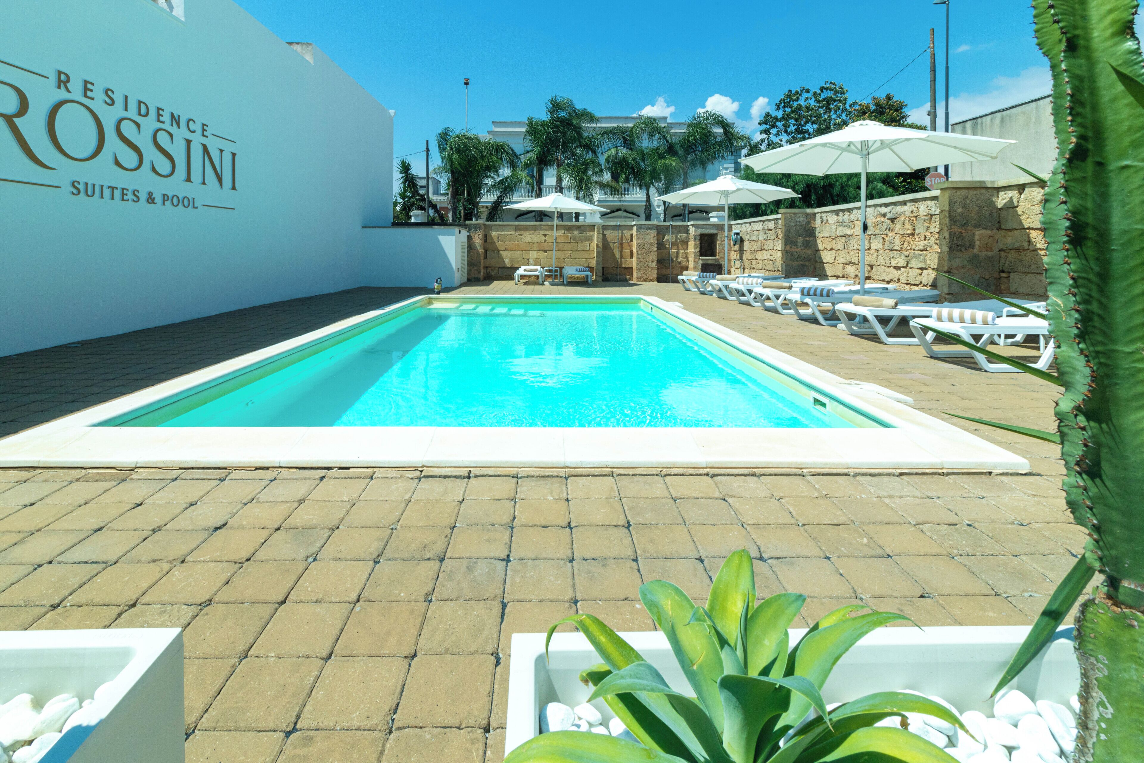 Una piscina con sillas de descanso y sombrillas, una pared con el texto 'Residence Rossini Suites & Pool', y un muro de piedra con palmeras al fondo.