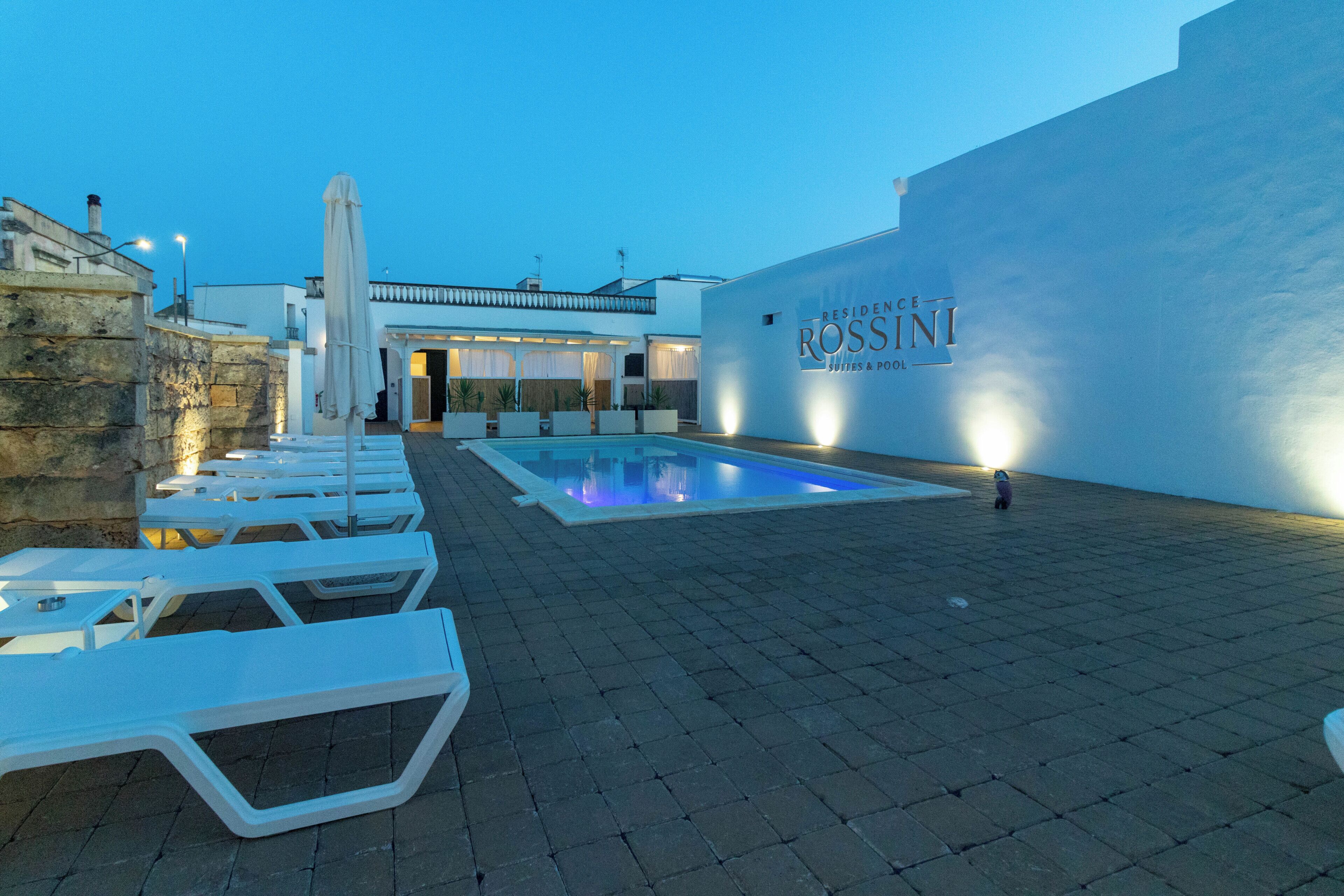 A pool area with lounge chairs, a stone wall, and a building with a sign reading 'RESIDENCE ROSSINI.'