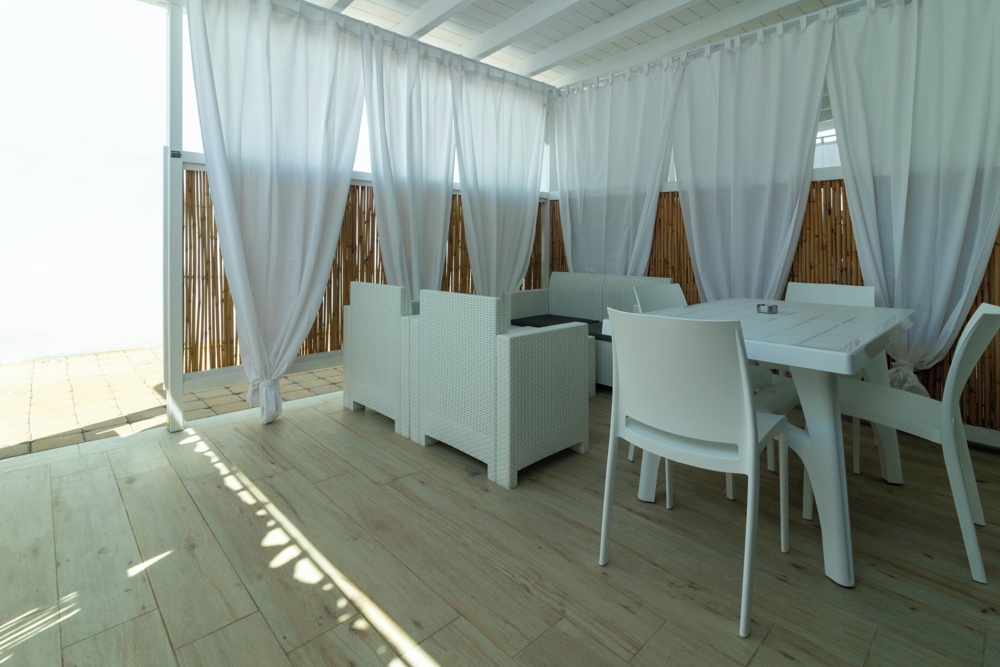 A room with a wooden floor, white furniture, and large windows with white curtains.