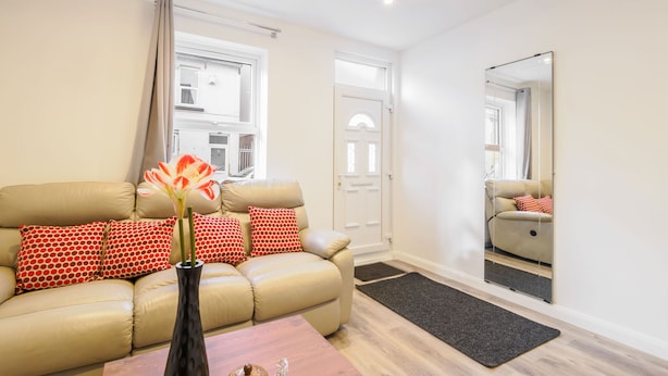 A living room with a beige sofa, red and white patterned pillows, a vase with flowers, a wooden coffee table, and a large mirror on the wall. - Host & Stay | Roach Street (Rochester)