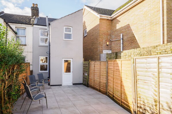 Un jardin à l’arrière d’une maison, avec une terrasse carrelée, une clôture en bois et une porte blanche.