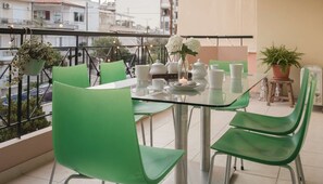 Un balcón con una mesa de vidrio dispuesta para el té, sillas verdes y plantas en macetas.