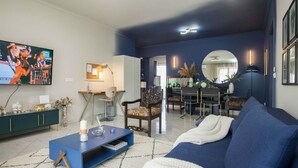 Un salon moderne avec un canapé bleu, une table basse et un téléviseur. On y trouve également un coin repas avec une table et des chaises, et une cuisine à l’arrière-plan.