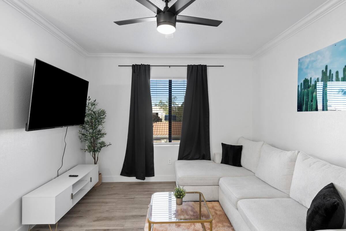 A modern living room with a flat-screen TV, a white sofa, a glass coffee table, and a ceiling fan.
