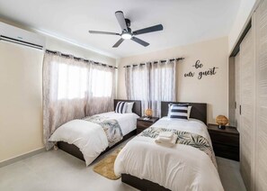 Quarto de hotel com duas camas, um ventilador de teto e um painel com a frase 'seja nosso convidado'.