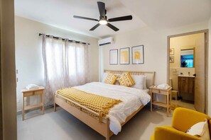 Ein Schlafzimmer mit einem Bett, einem Deckenventilator, einem Nachttisch, einem Sessel und einem durch eine offene Tür einsehbaren Badezimmer.